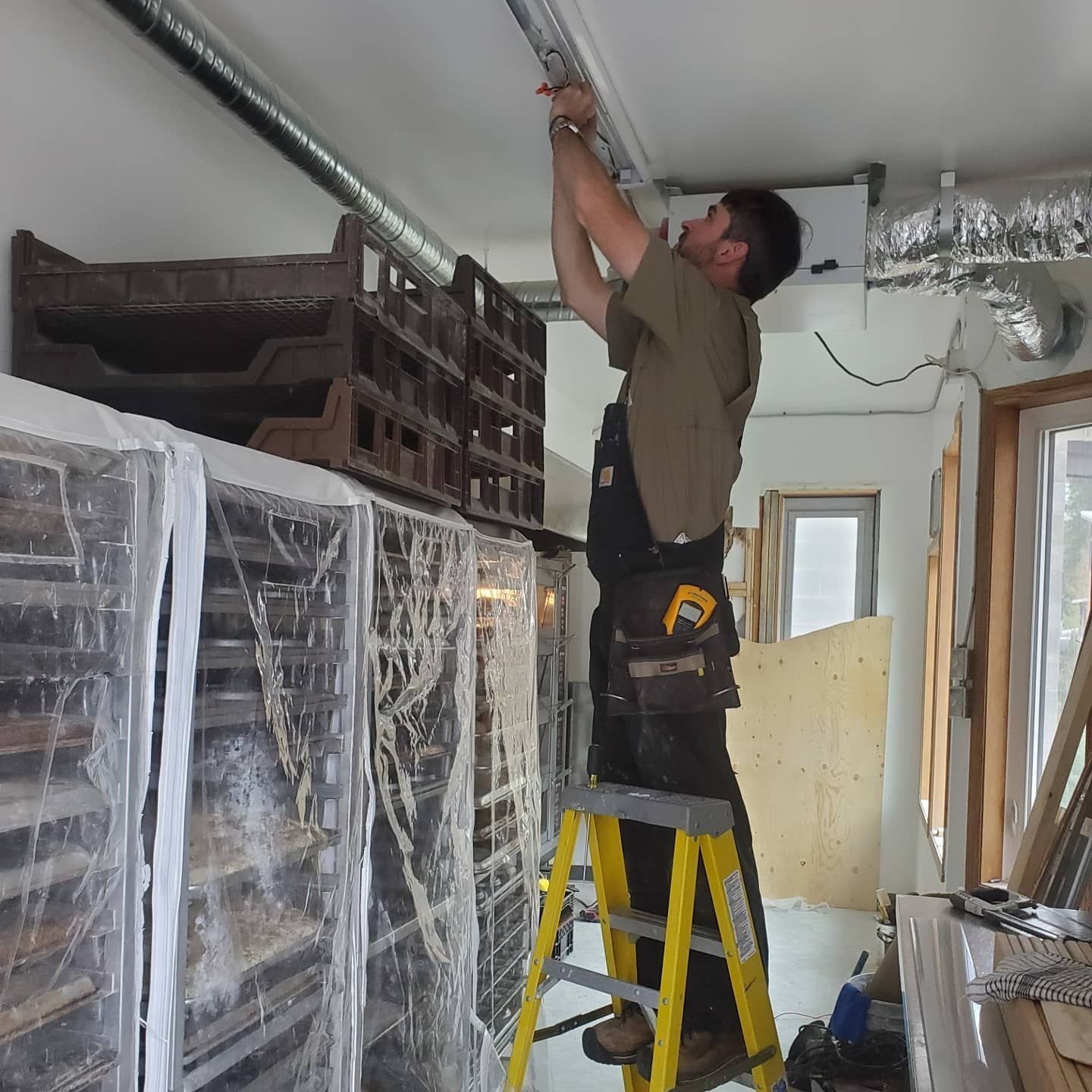 Worker on a yellow ladder installing ductwork in a room under construction.