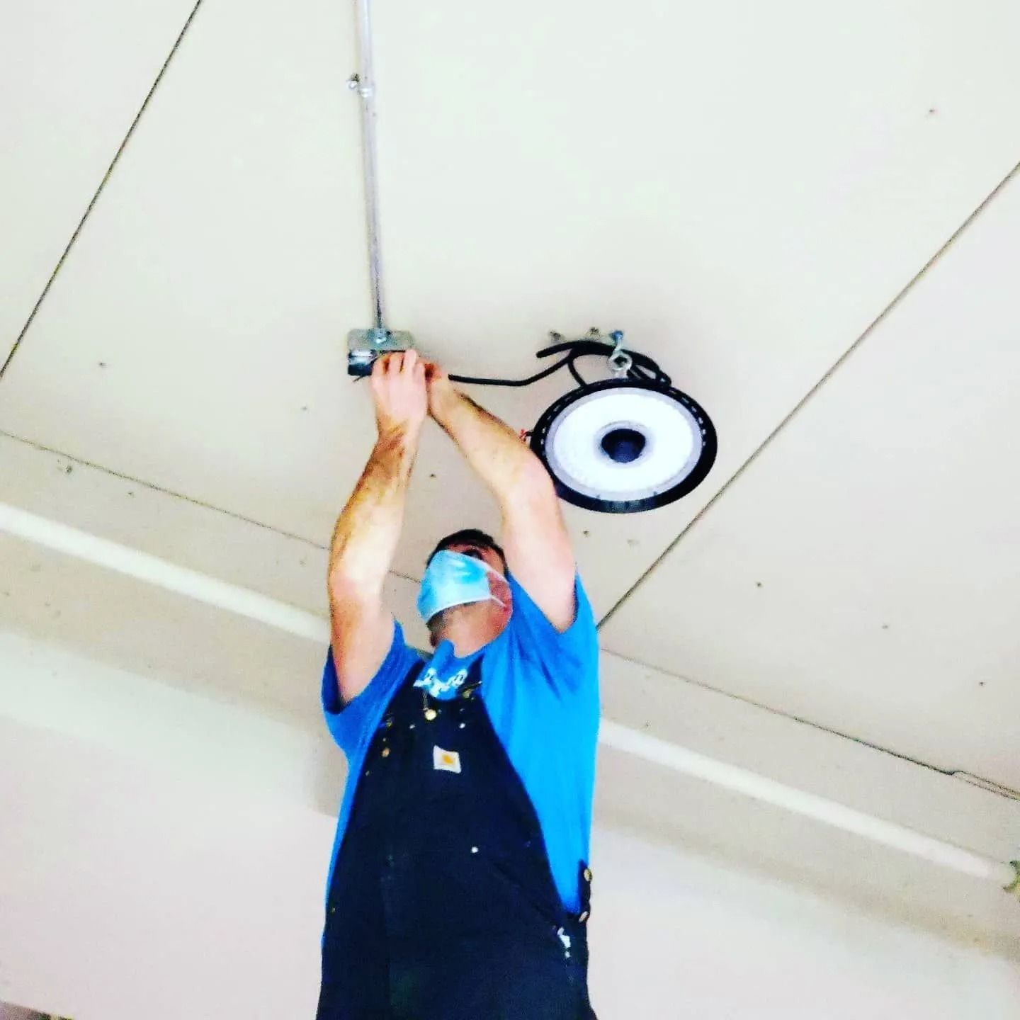 Worker in blue shirt and mask installing a ceiling light fixture with raised arms
