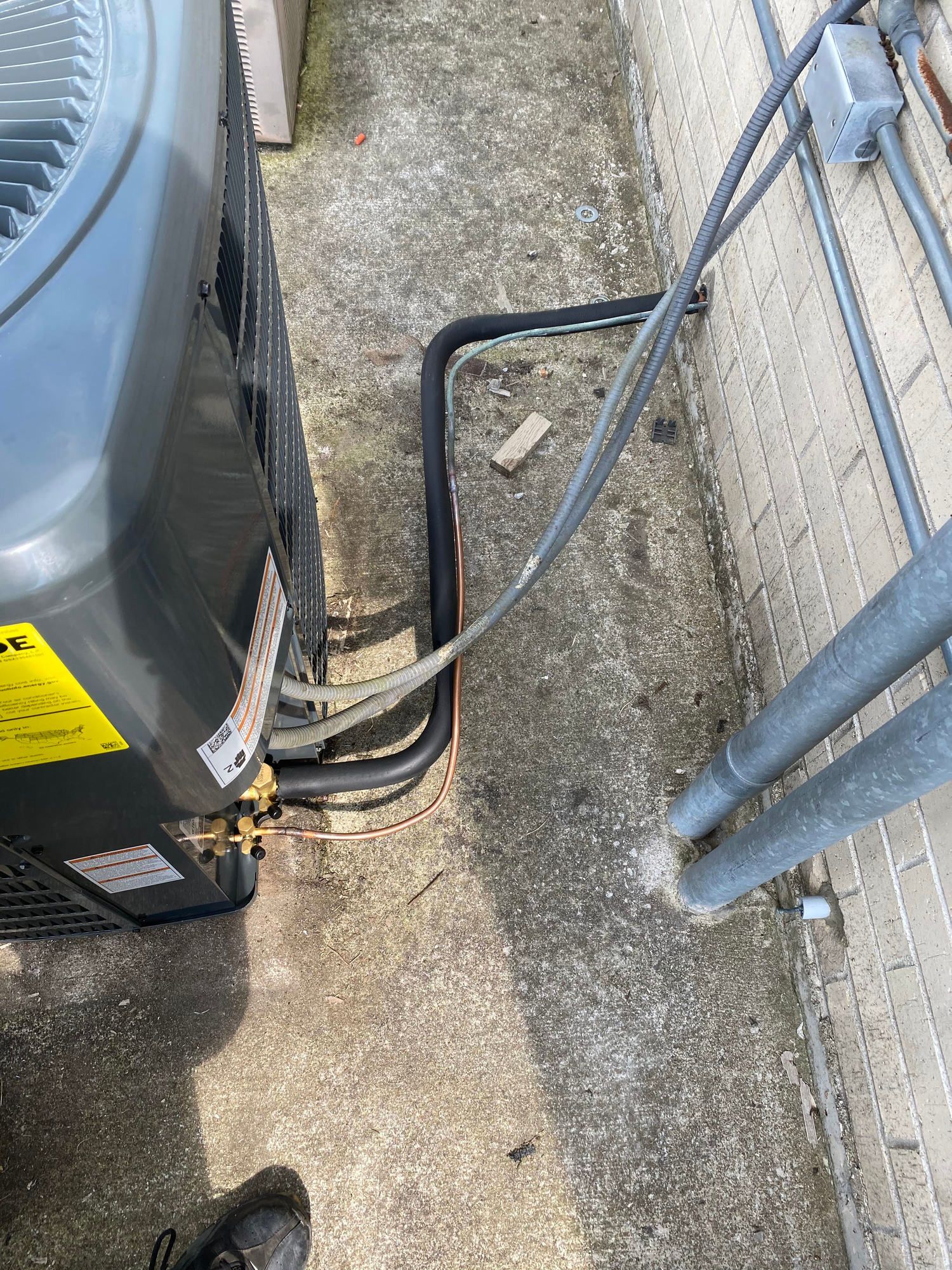 A gray air conditioner is sitting on the sidewalk next to a brick wall.