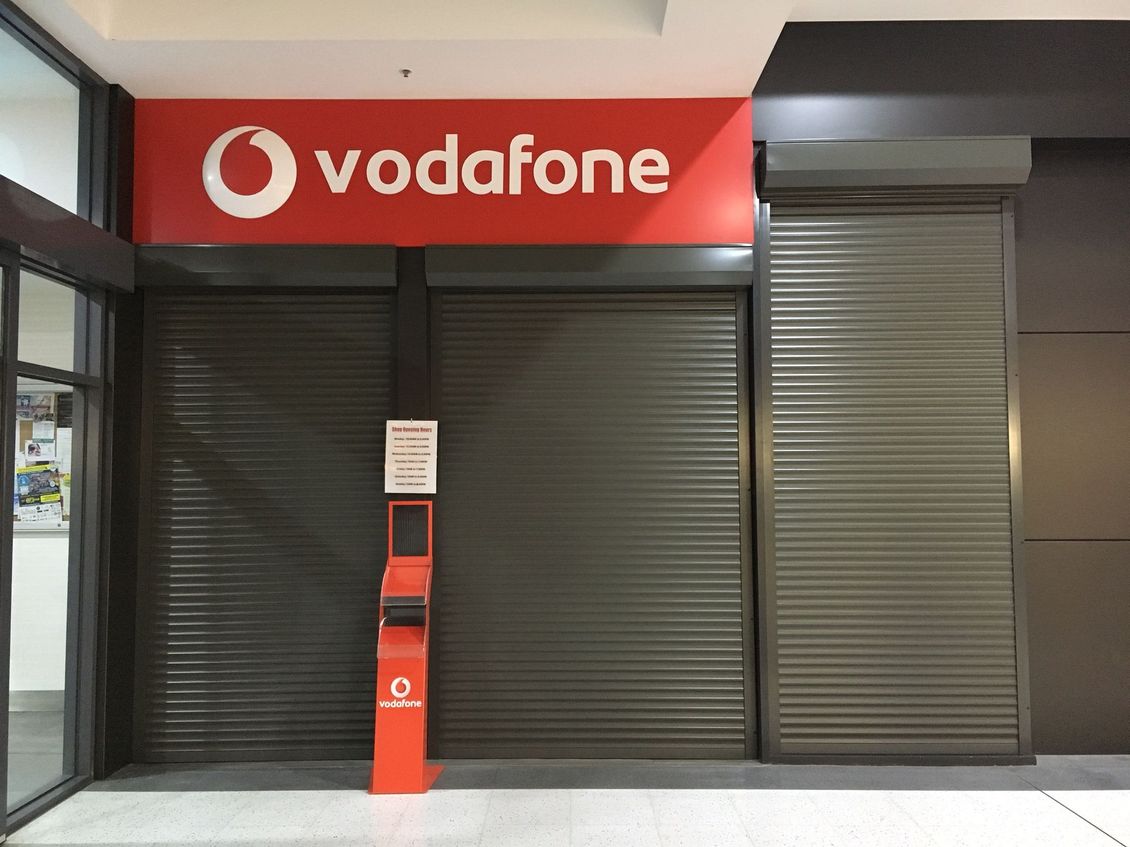 A closed, grey metal rolling shutter door installed in a white wall next to a large multi-pane window and a fire extinguisher.