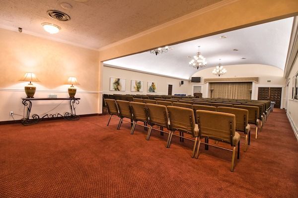 A funeral home chapel with rows of chairs facing a stage, red carpet, cream walls, lamps, and art.