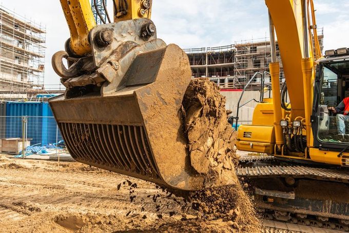Yellow excavator dumping dirt at a construction site, with buildings and scaffolding in the background.