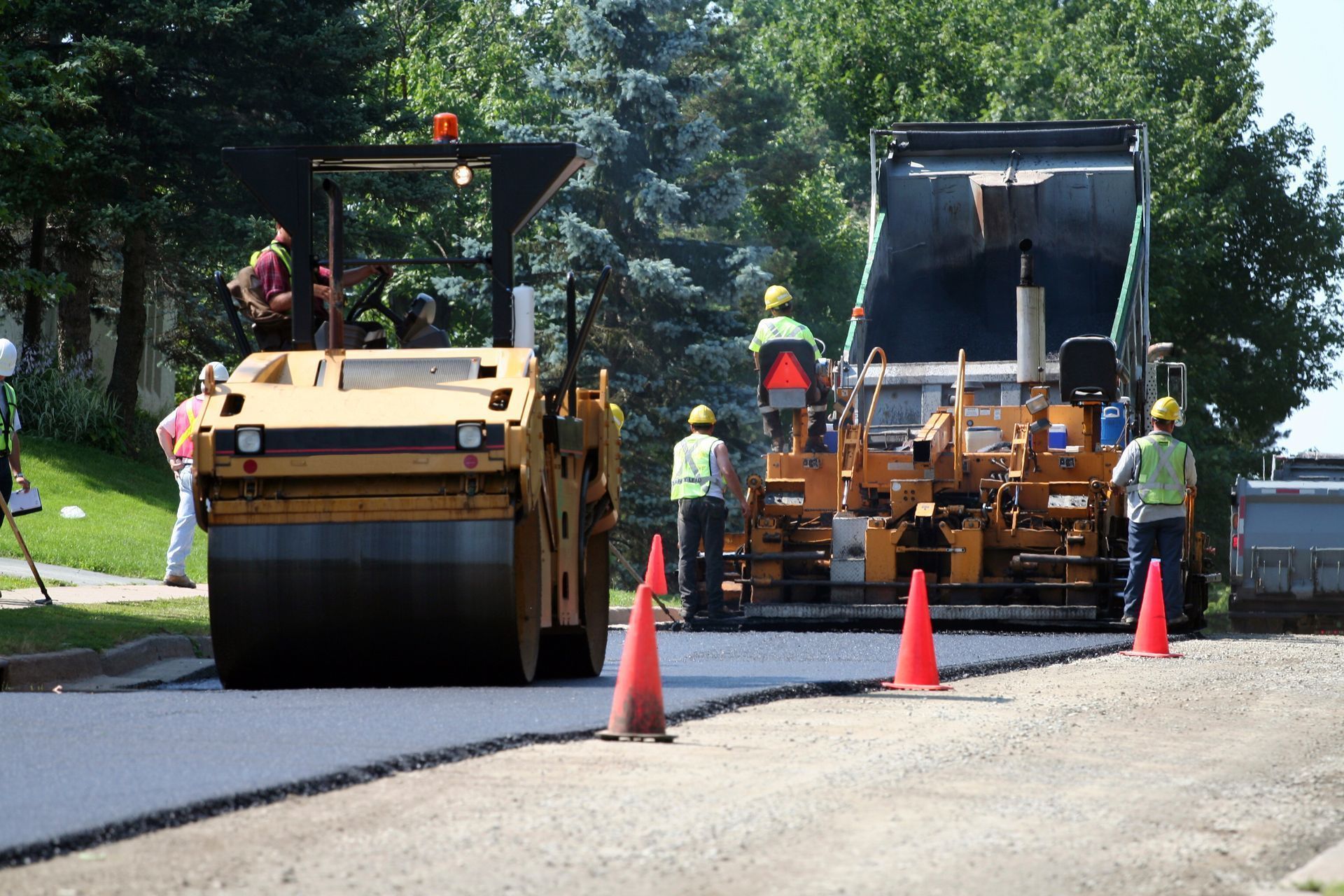 Road construction: Asphalt paving with heavy machinery, workers in safety vests, cones set out.