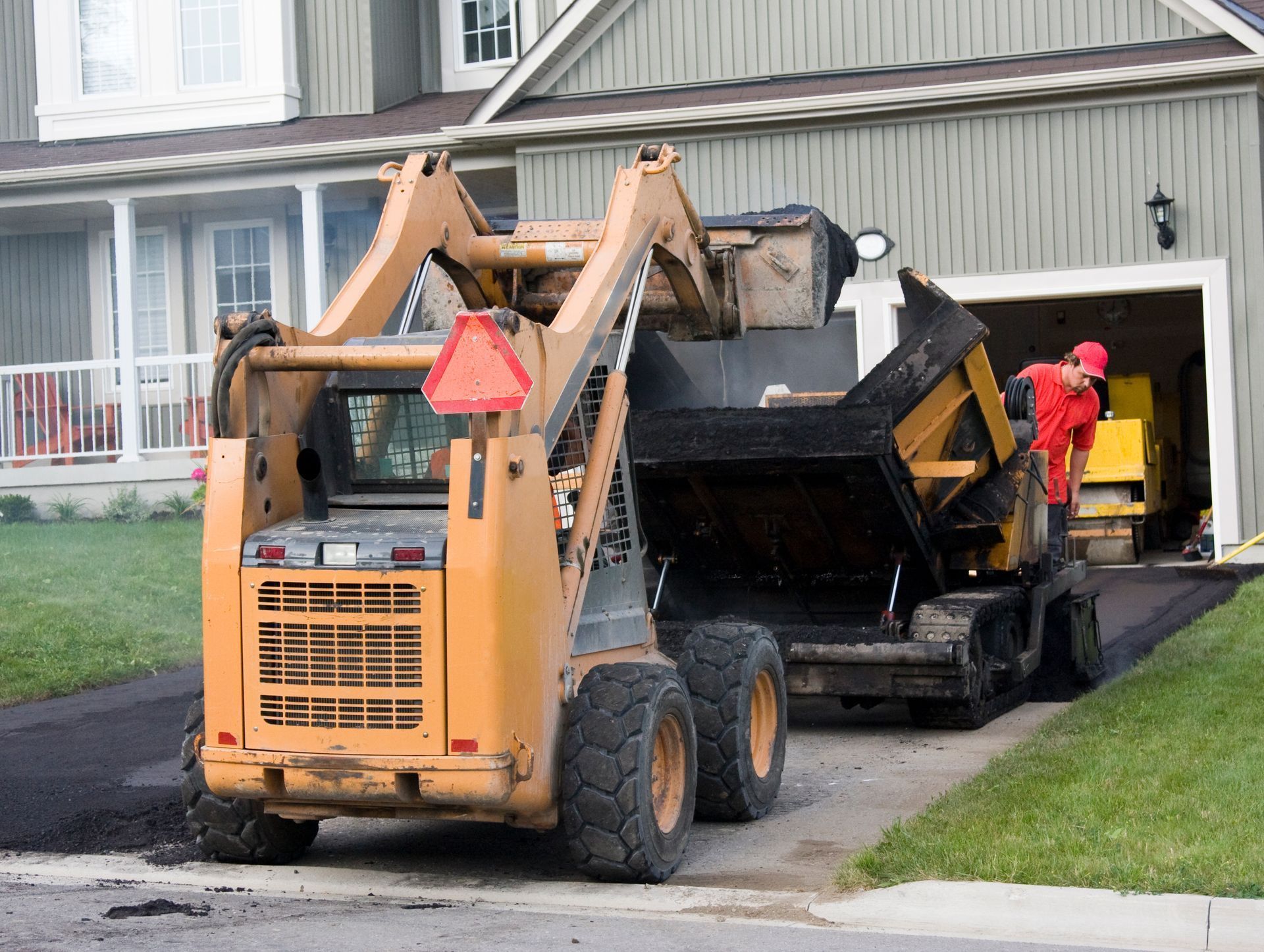 A skid steer loader unloads asphalt onto a driveway near a house, while a worker in red stands in the garage.