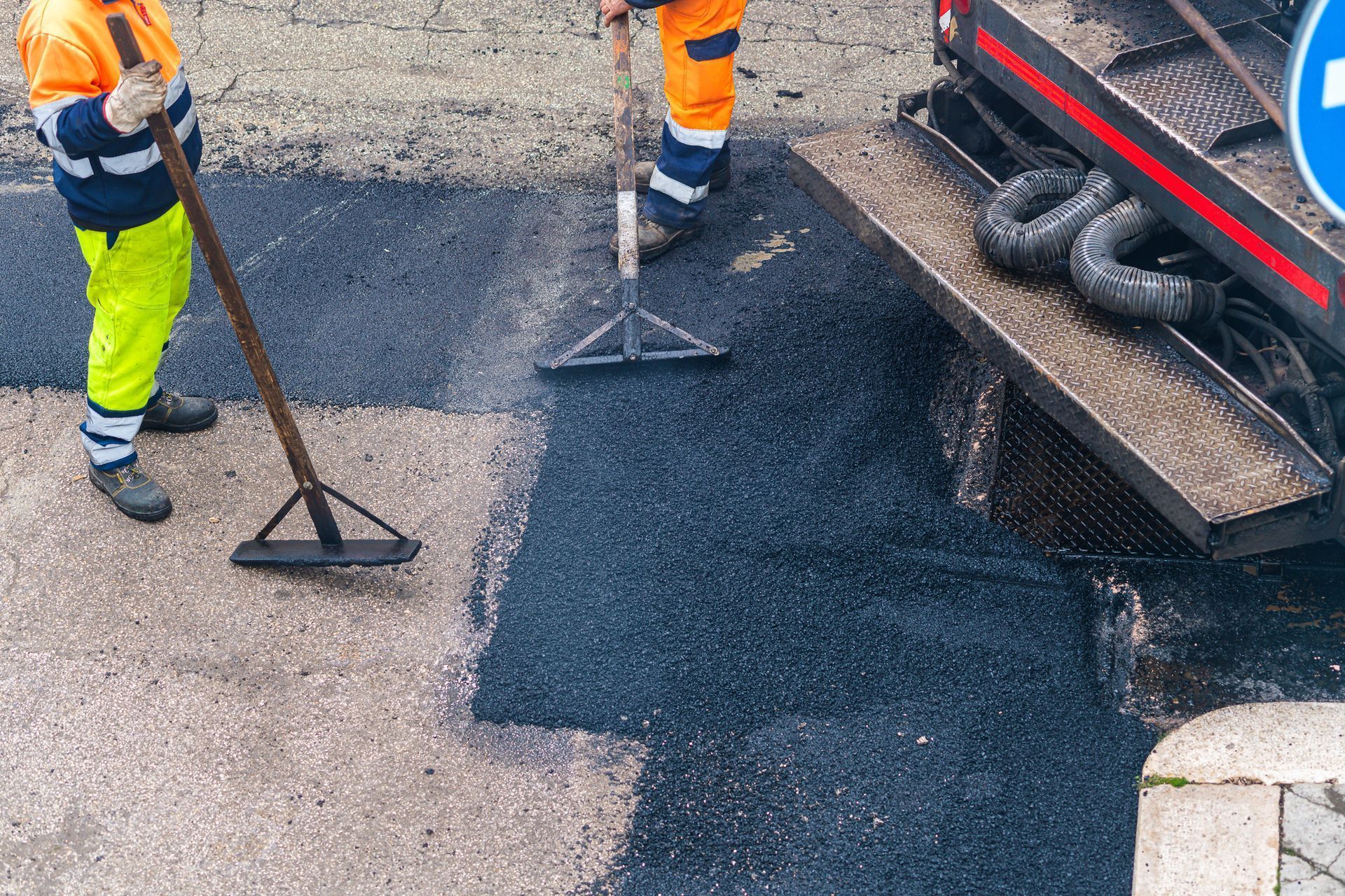 Two road workers in safety gear laying asphalt on a street from a truck. One uses a rake, the other a shovel.