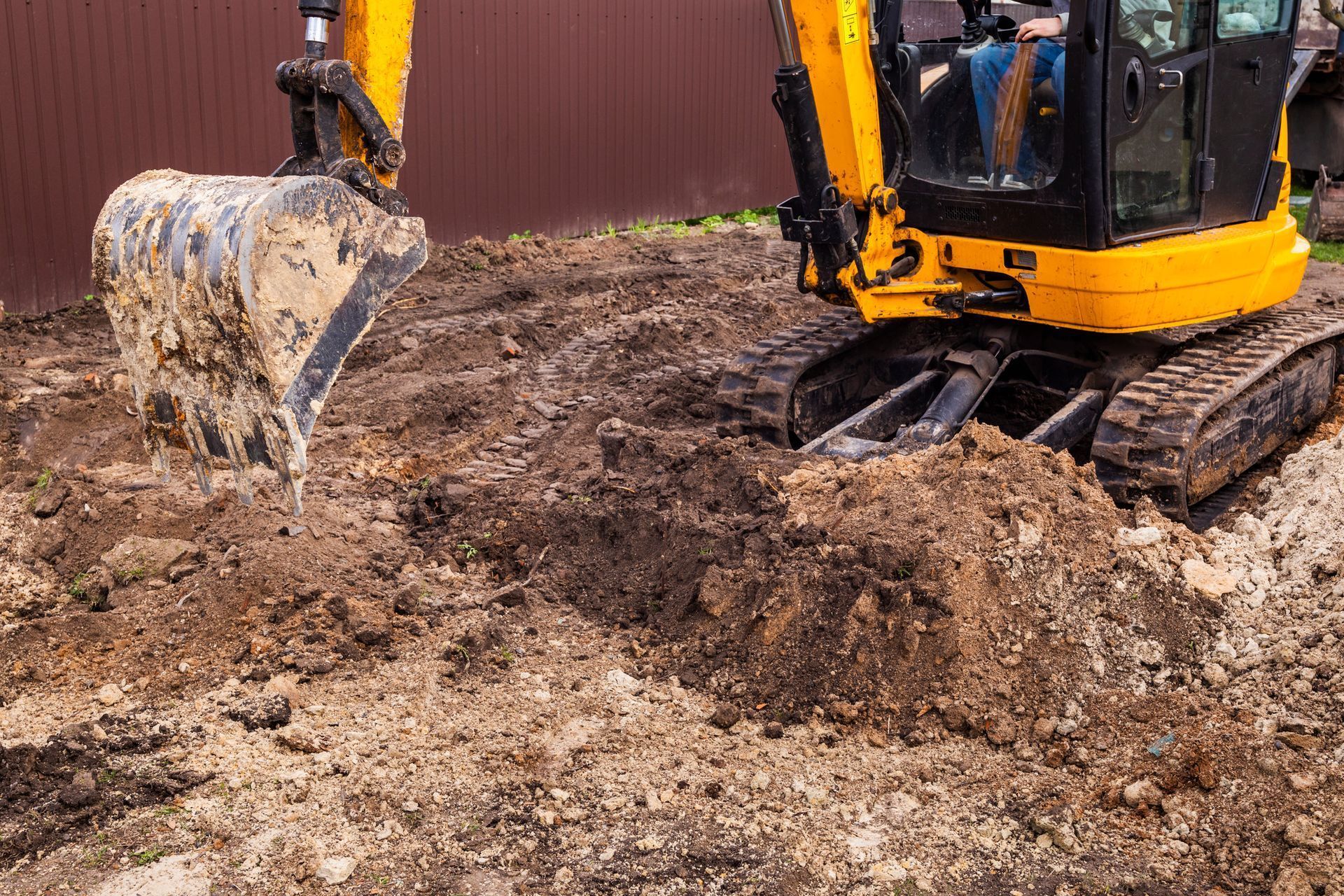 Yellow excavator digging a trench in brown soil, near a brown fence.