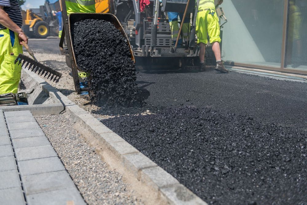 Workers paving a road with hot asphalt, pouring from a machine. They are wearing safety vests and using rakes.