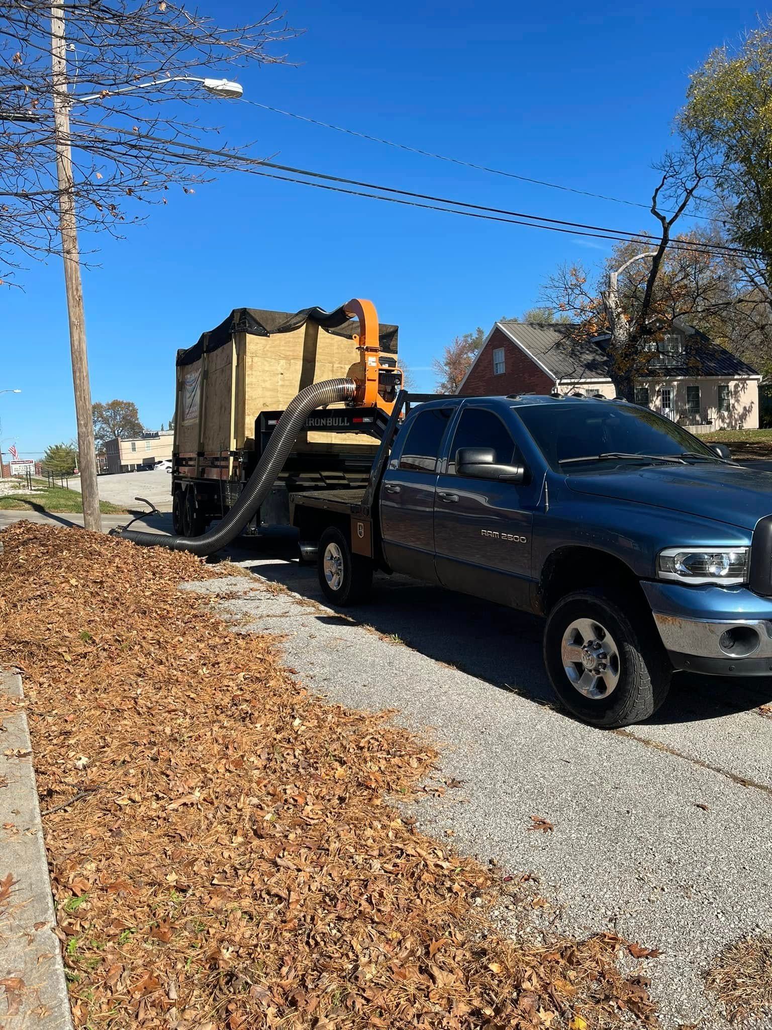 Blue pickup truck with leaf vacuum trailer collecting leaves on a street.