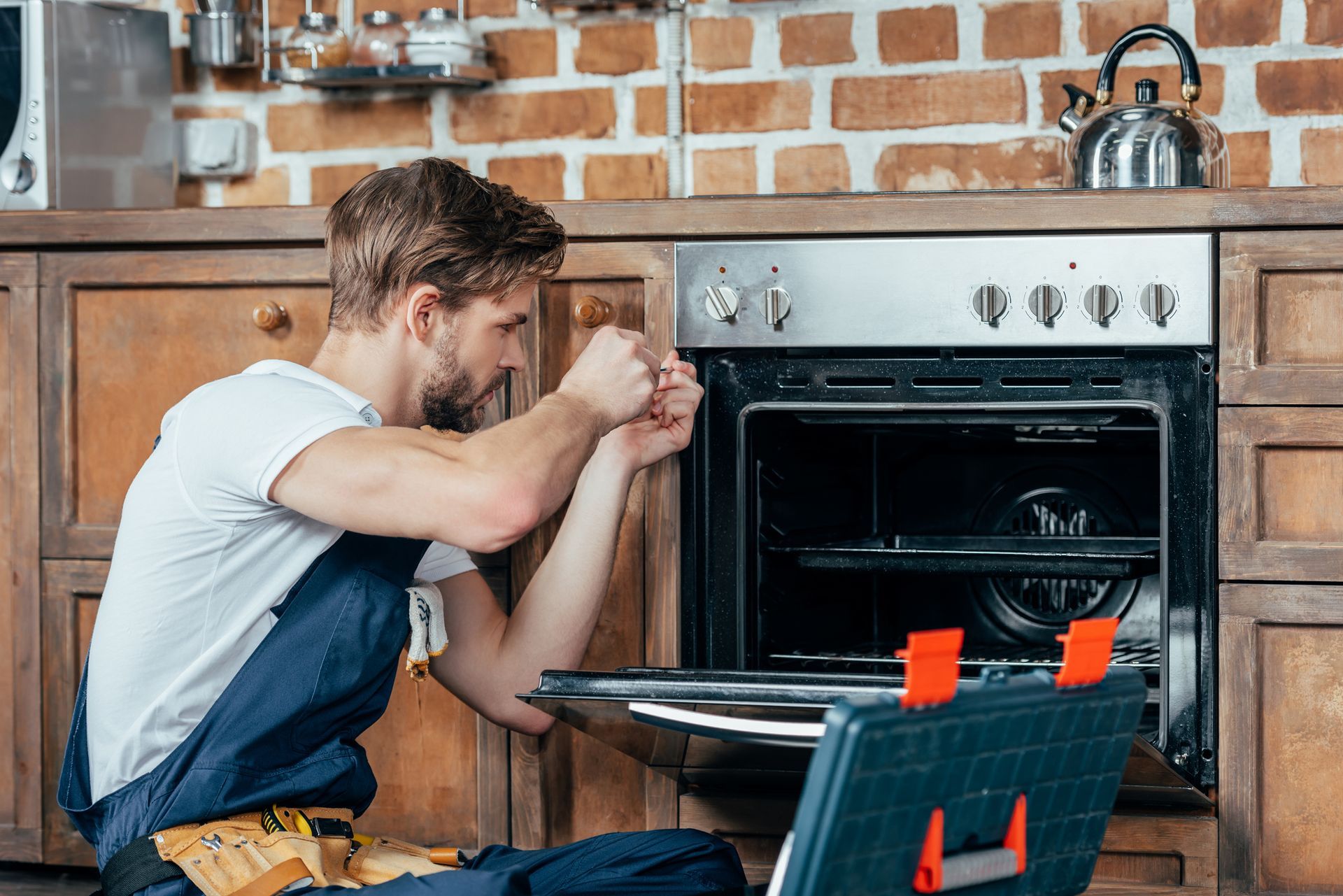 Young technician in protective workwear, performing oven range repair in a residential kitchen.