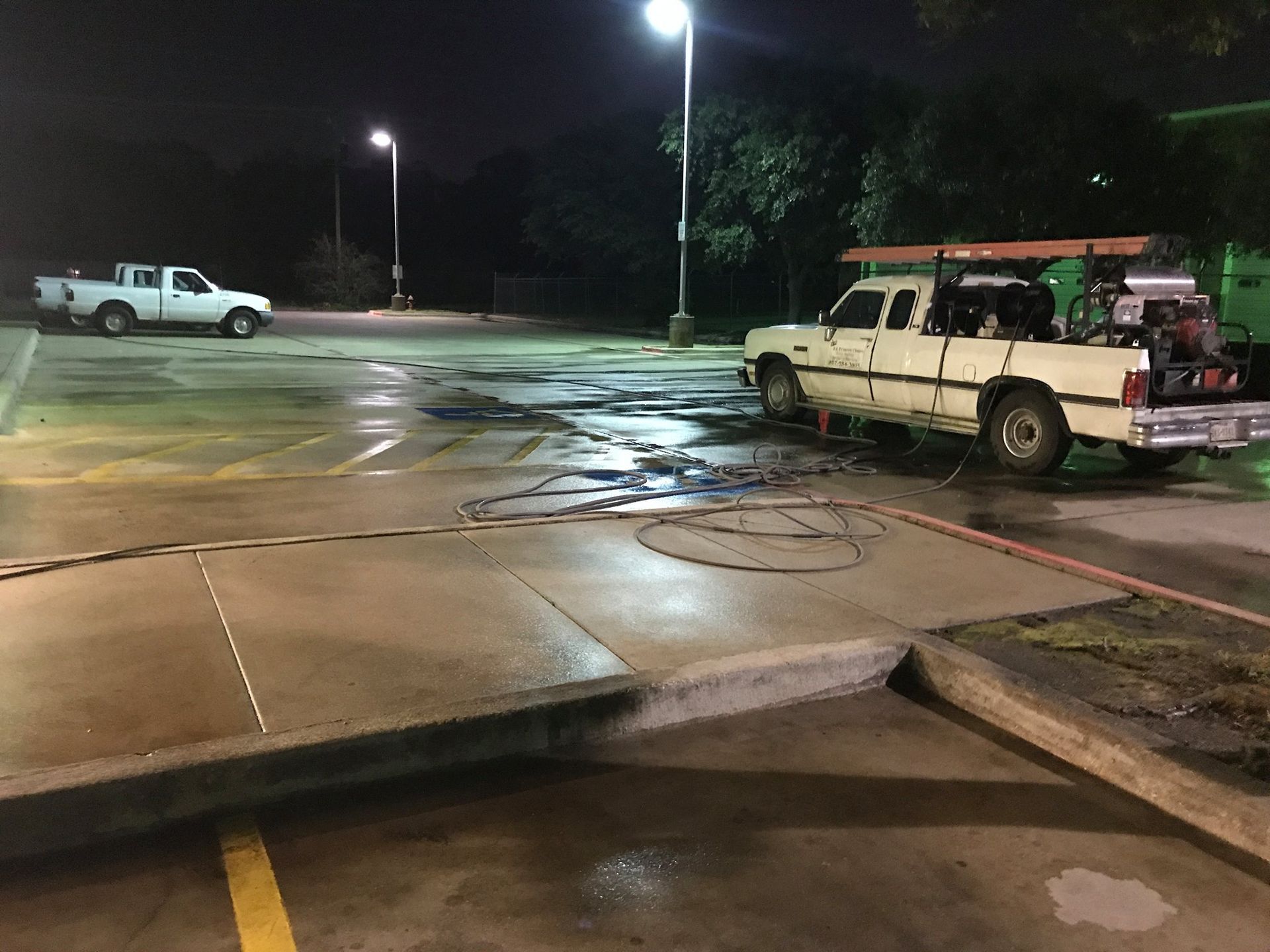 Two work trucks in an empty parking lot at night. Hoses are strewn across the concrete.