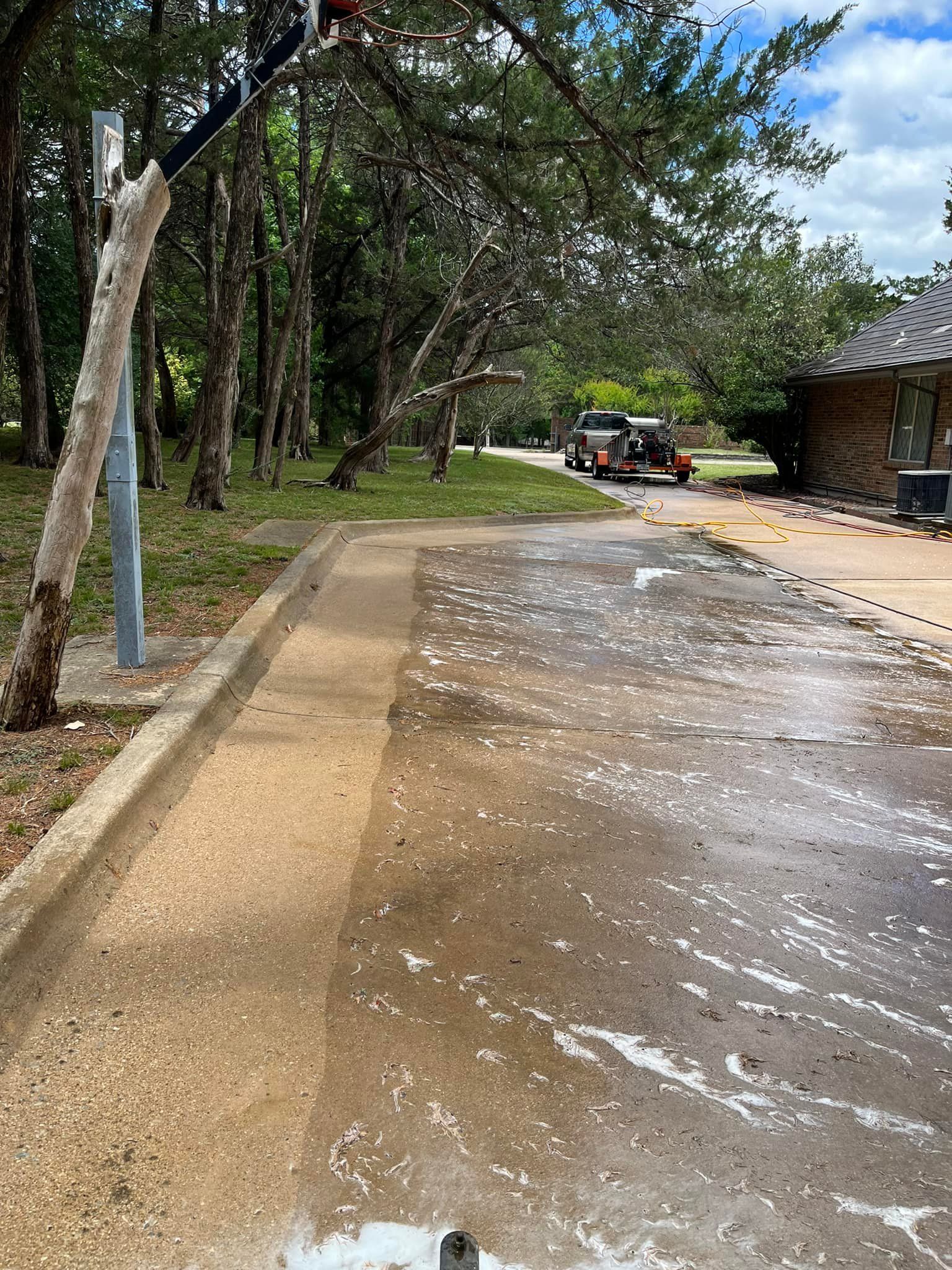 A concrete basketball court being power-washed outside. Trees and a building are visible in the background.