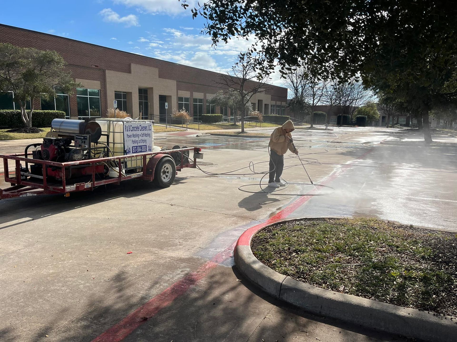 Person pressure washing a parking area with a trailer-mounted machine. Building and trees in the background.