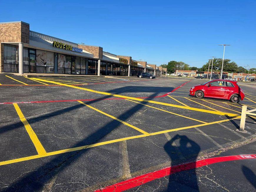 A red car parked in front of a strip mall with yellow and red painted parking lines on a sunny day.