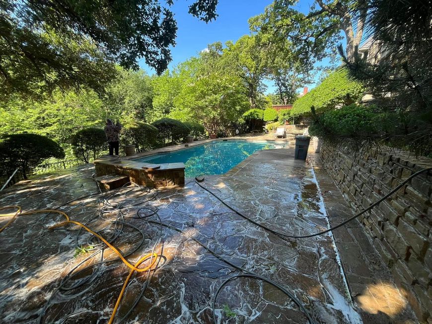 Poolside cleaning: A person pressure washes a stone patio, spraying water. Pool and greenery in background.