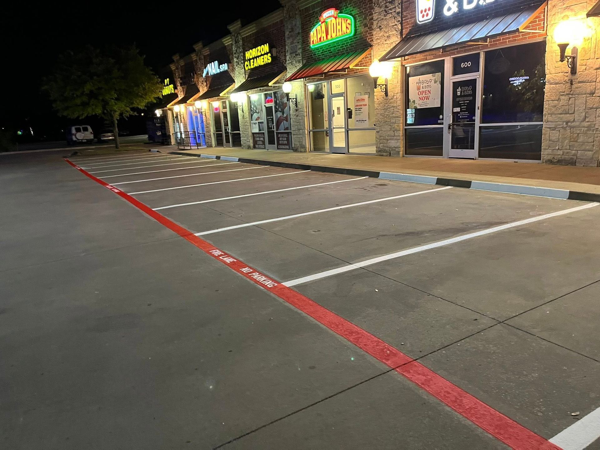 Nighttime view of an empty parking lot in front of a strip mall with illuminated storefronts and parking space lines.