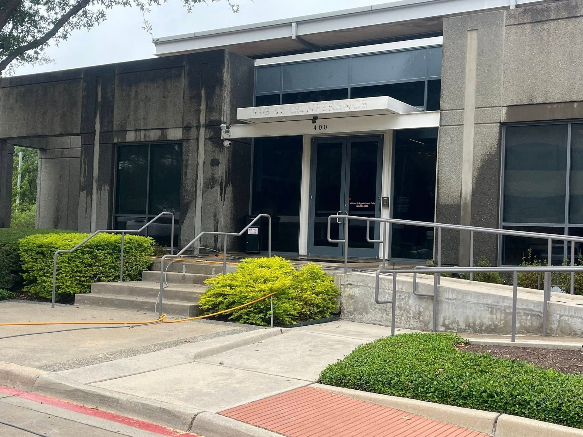 Gray building with dark windows, a ramp, and stairs leading to the entrance. Green bushes line the front.