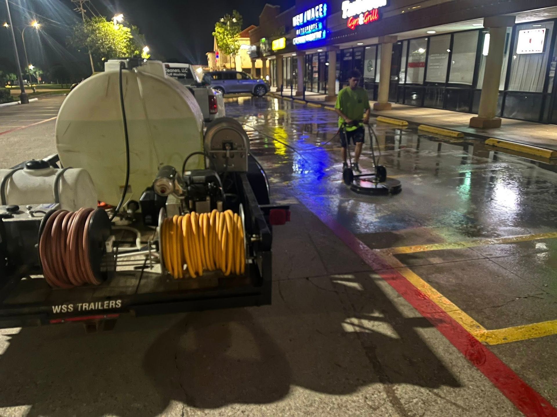 Person using a power washer to clean a wet parking lot at night. A trailer with tanks and hoses is visible.