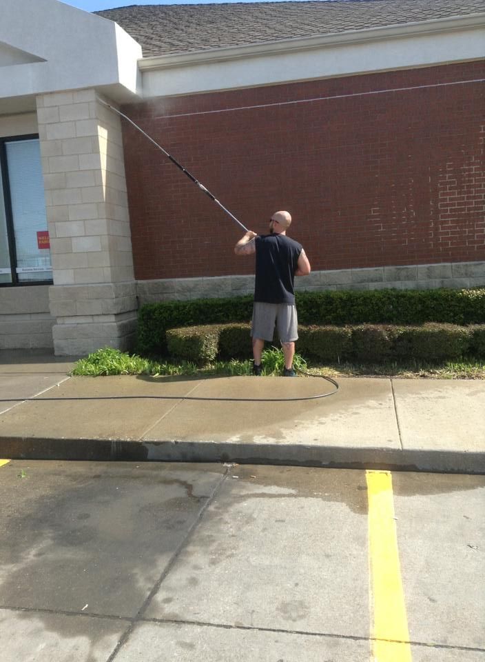 Man pressure washing a brick wall outside a building, spraying water with a long handled wand.