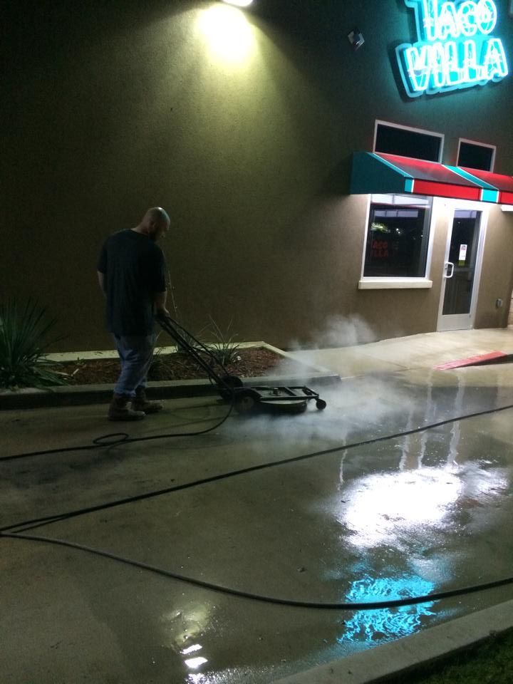 A man pressure washes the concrete in front of Taco Villa at night; steam rises from the wet ground.