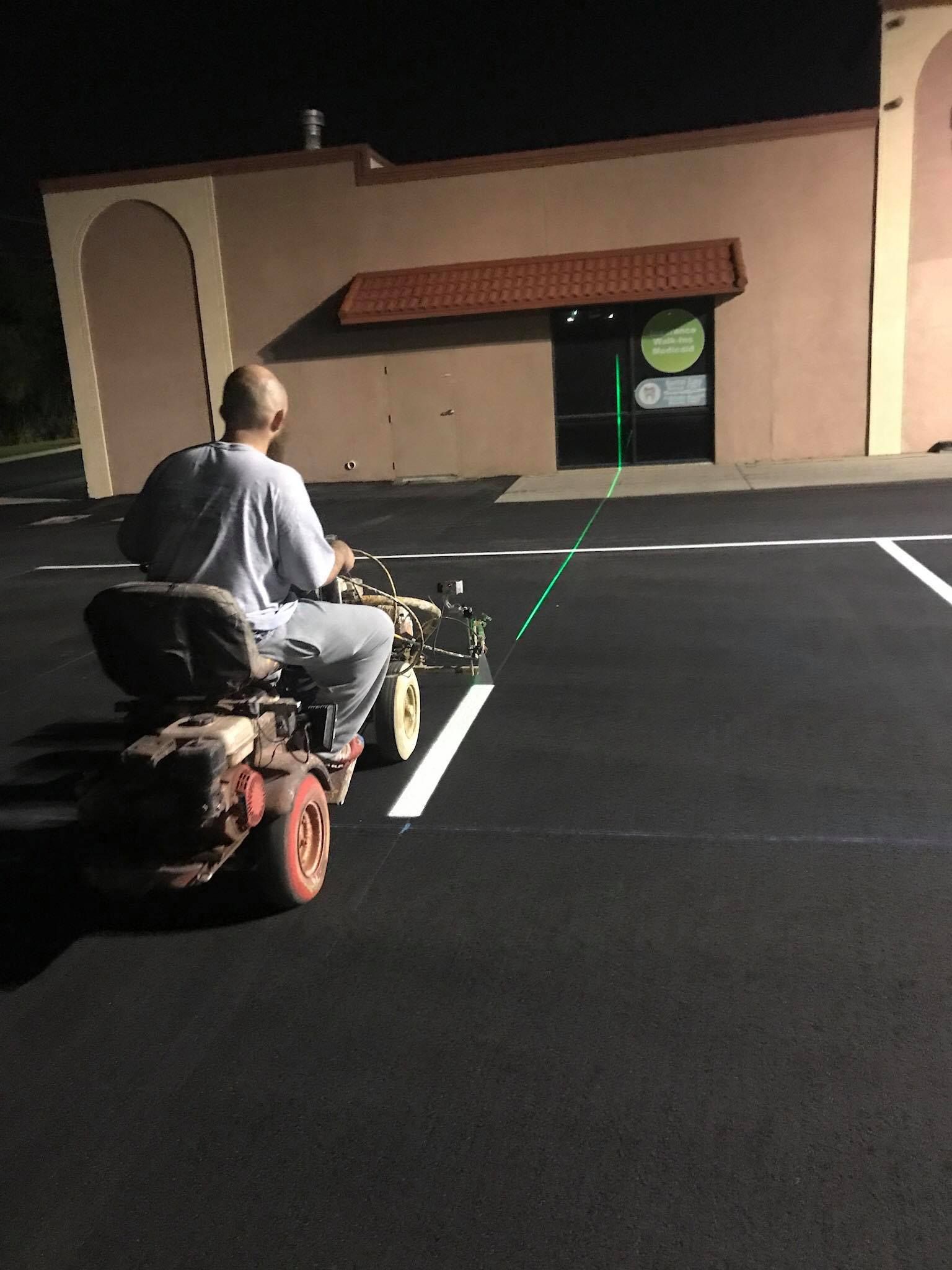 Man on parking lot striping machine at night. Dark asphalt, white lines. Building in background.