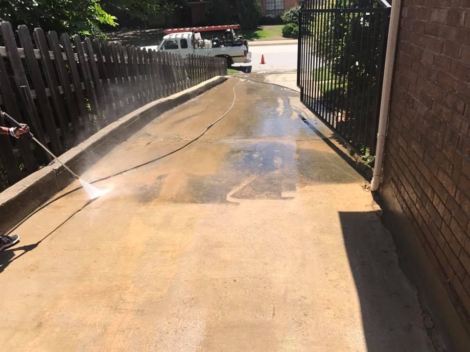 Person pressure washing a concrete driveway next to a wooden fence and a brick wall.