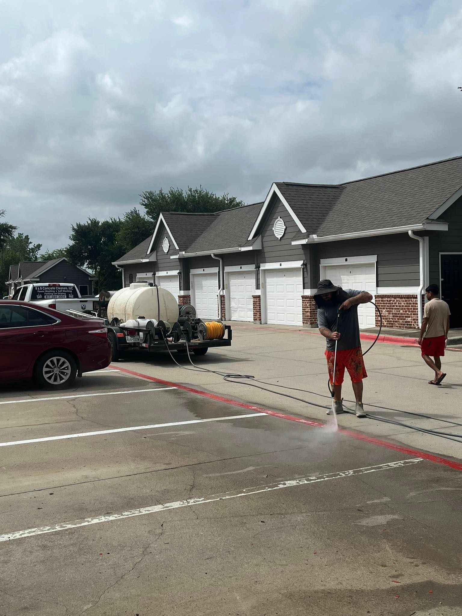 Person pressure washing pavement near red car and buildings.