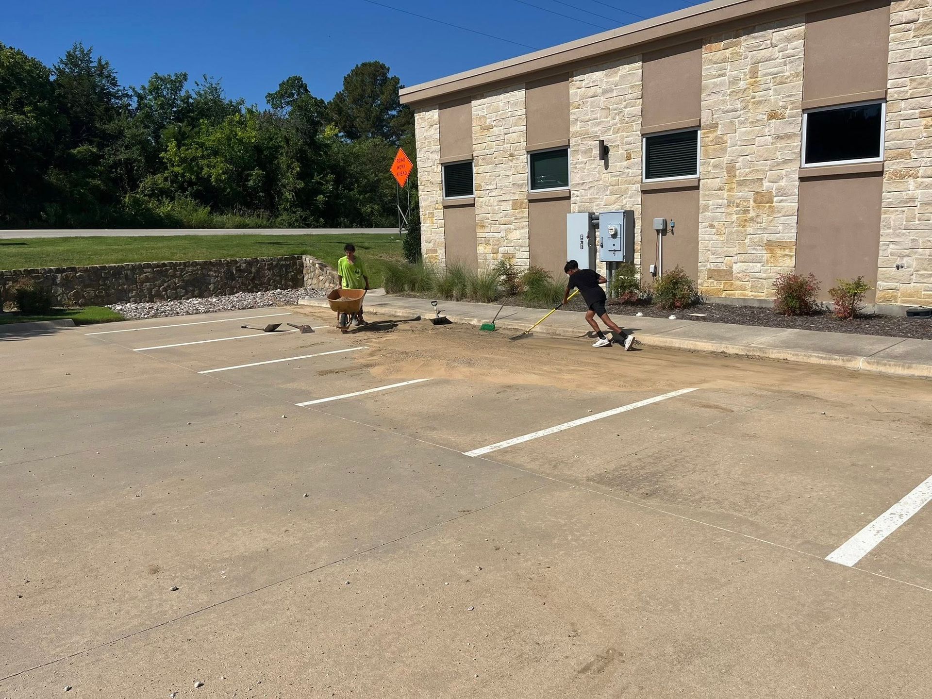 Two workers spread gravel in a parking lot. Beige building with stone facade. Sunny day.