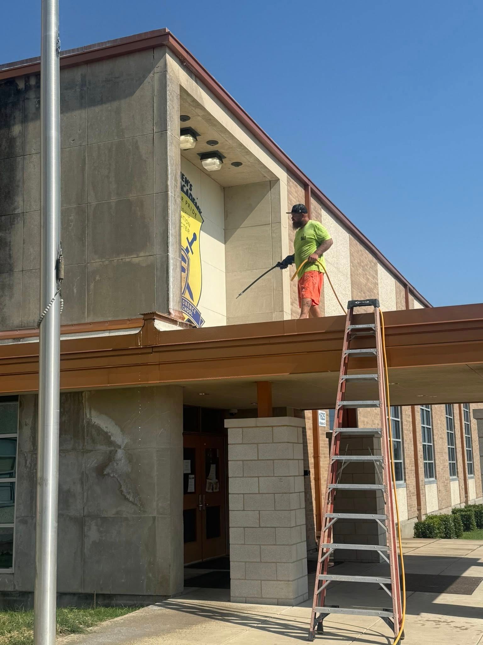 Man on ladder power washes exterior wall of building.