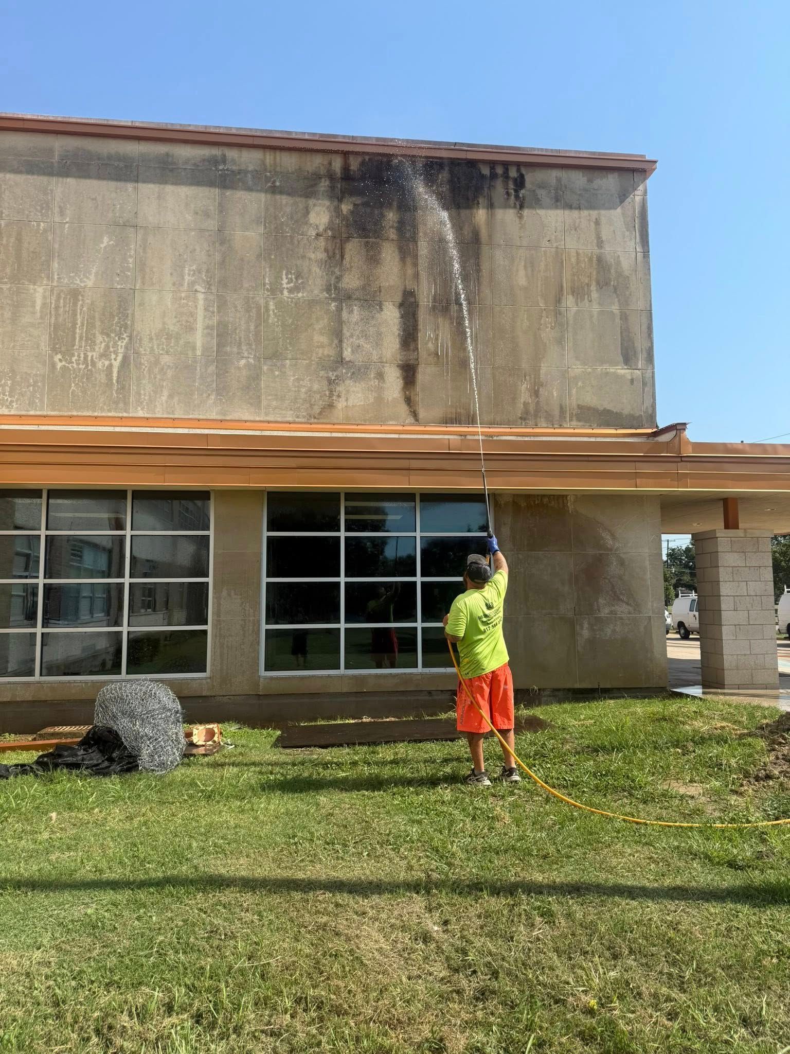 Person in neon shirt power washing a building's weathered exterior, near windows, under a blue sky.