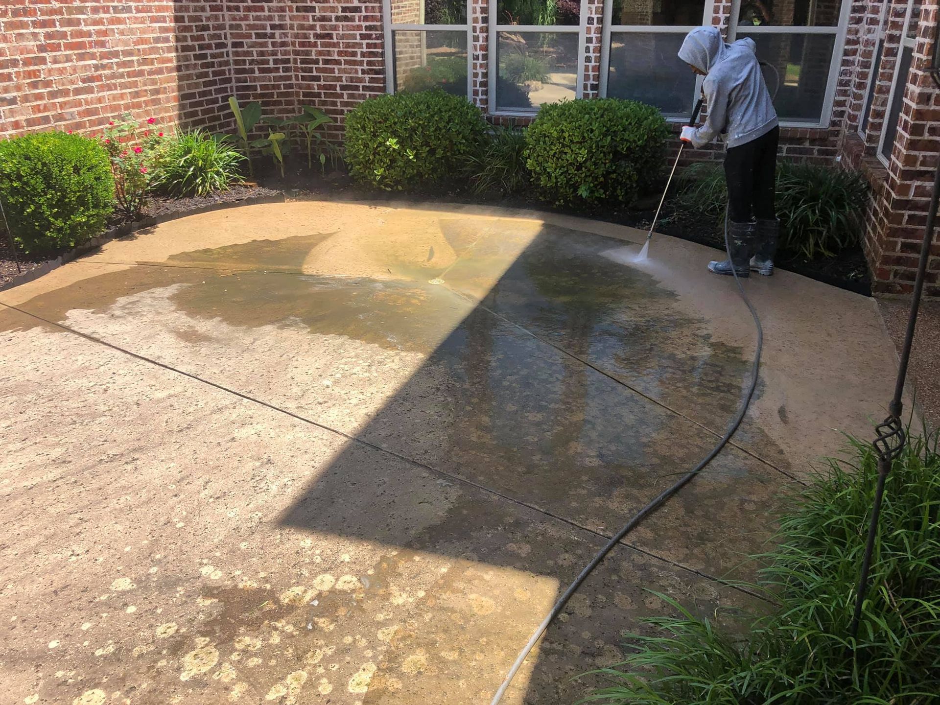 Person power washing a concrete patio next to a brick house and garden.