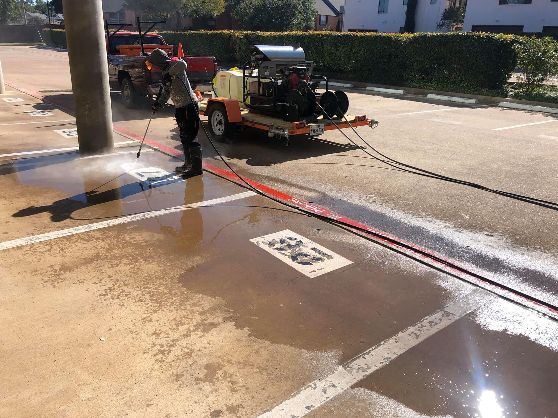 Person power washing a parking lot; trailer with cleaning equipment. Sunny day.