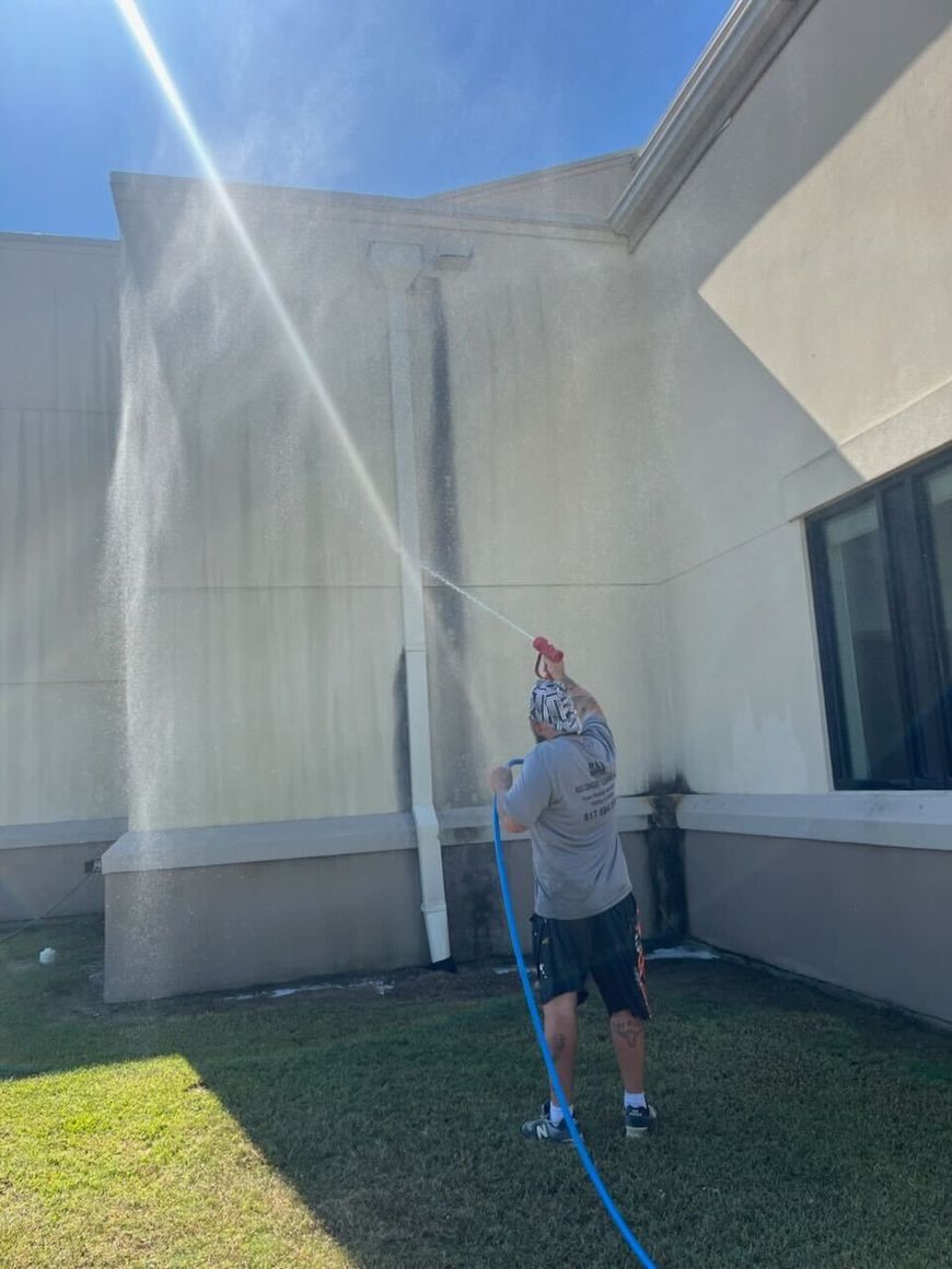 Person washing exterior wall of a building with a hose on a sunny day.