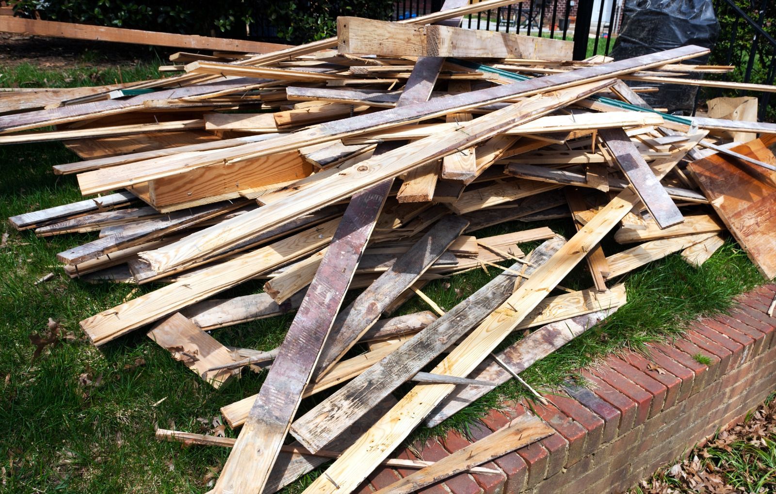 Pile of splintered wood planks and scraps on green grass next to a brick wall.