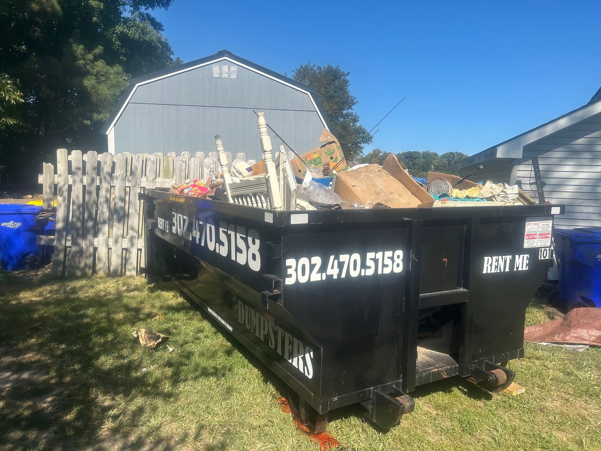 A black dumpster overflowing with debris sits on grass in front of a wooden fence and blue-grey building on a sunny day.