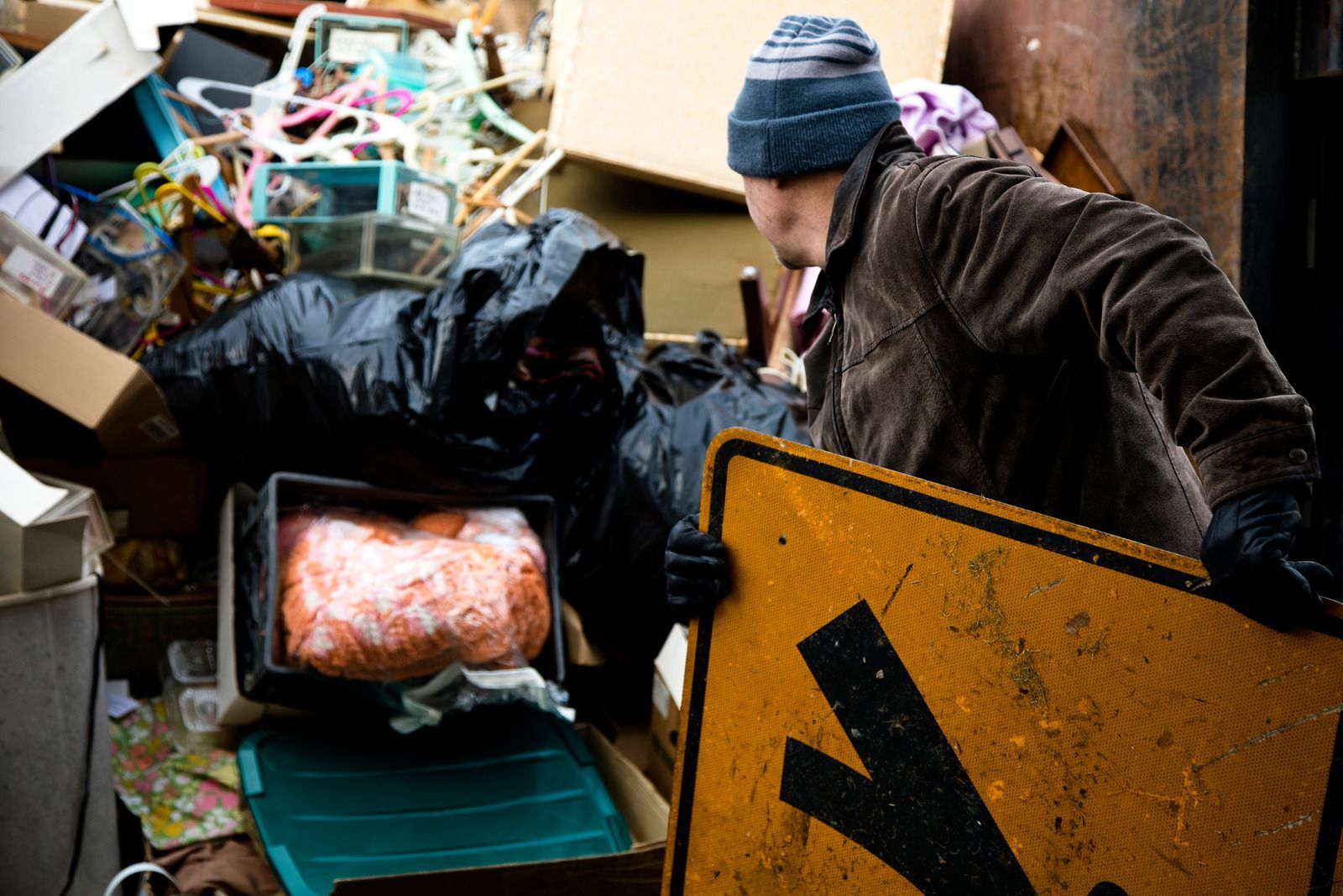 Man in coat rummages through a pile of trash, including a yellow road sign.