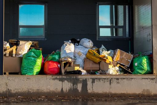 Pile of trash bags and boxes on a concrete surface against a building with windows; various colors.