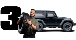Man holding a spare tire beside a black off-road SUV with a large number 3 in the background | 106 St Tire and Wheel