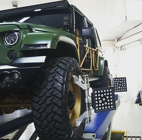 Green off-road truck on a lift in a garage, with large tires and exposed suspension modifications. | 106 St Tire and Wheel