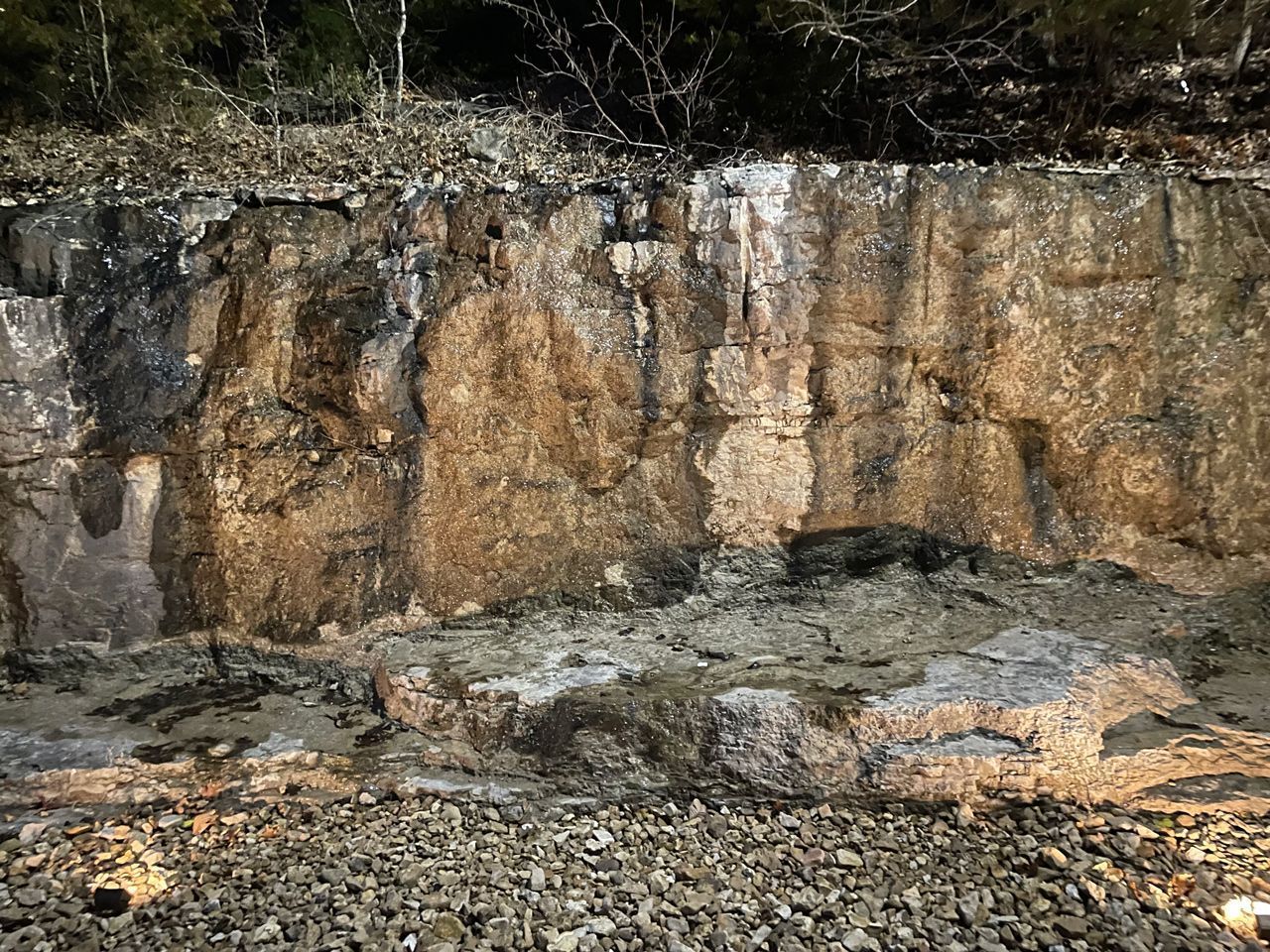 A large rock wall is surrounded by gravel and trees.