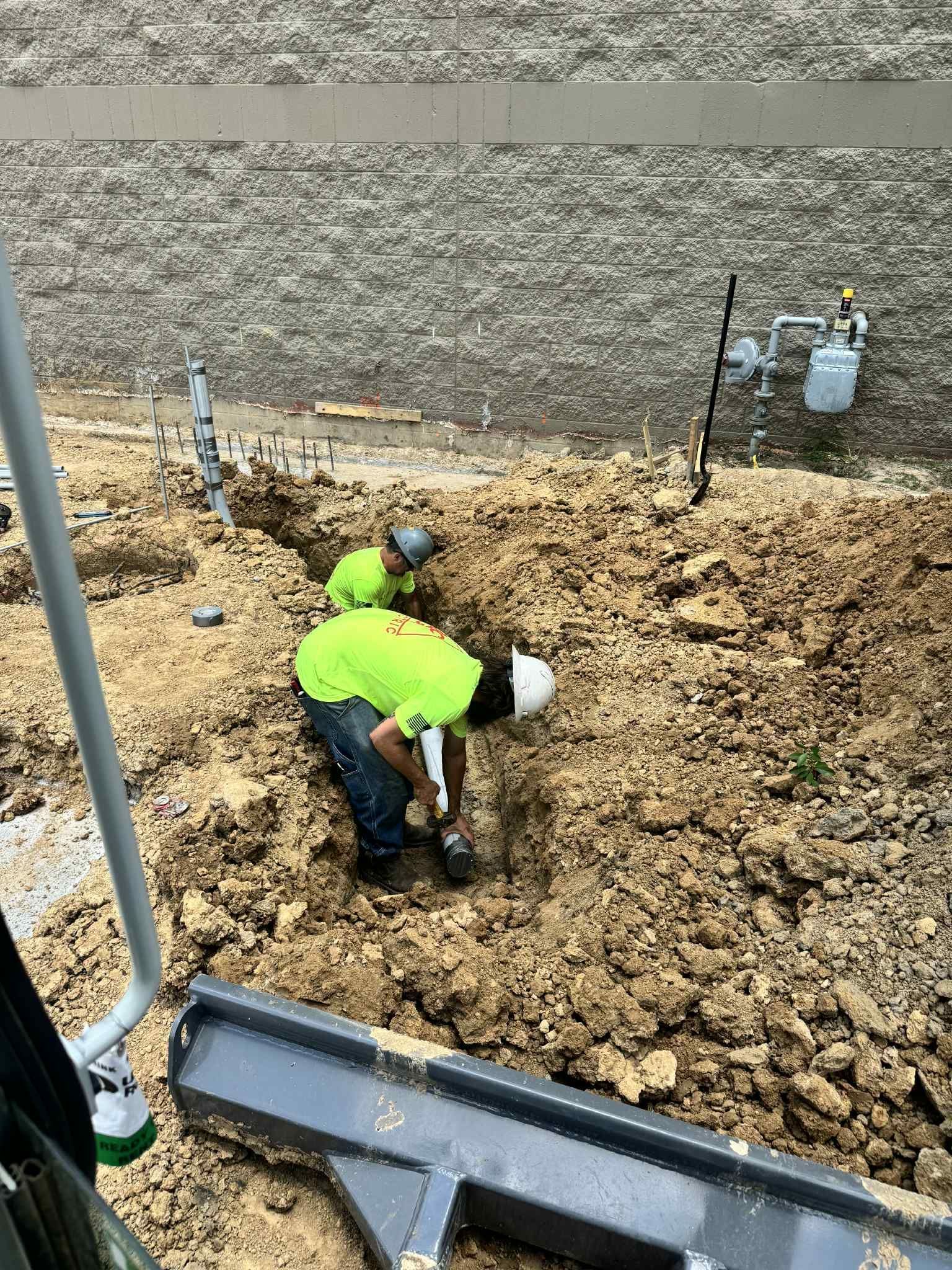 Two workers in safety vests dig a trench outdoors near a building.