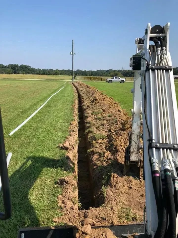 Excavator digging a trench on a grassy field, with a white utility line marked with paint.