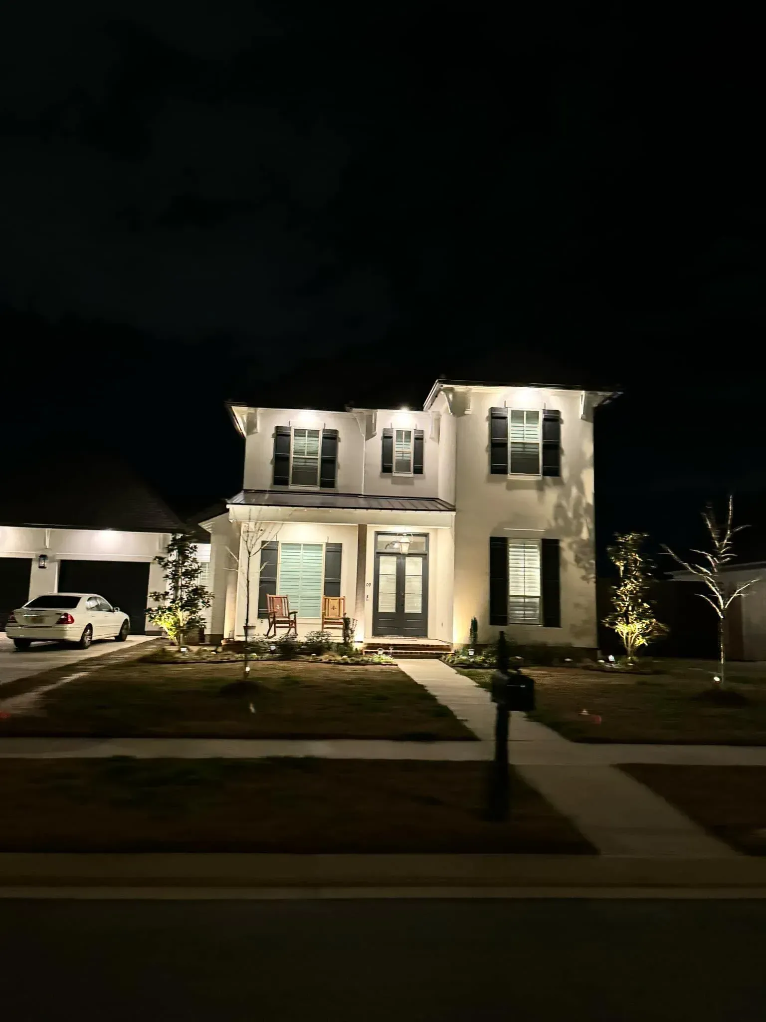 Two-story white house with black shutters, lit up at night. A car is parked in the driveway.