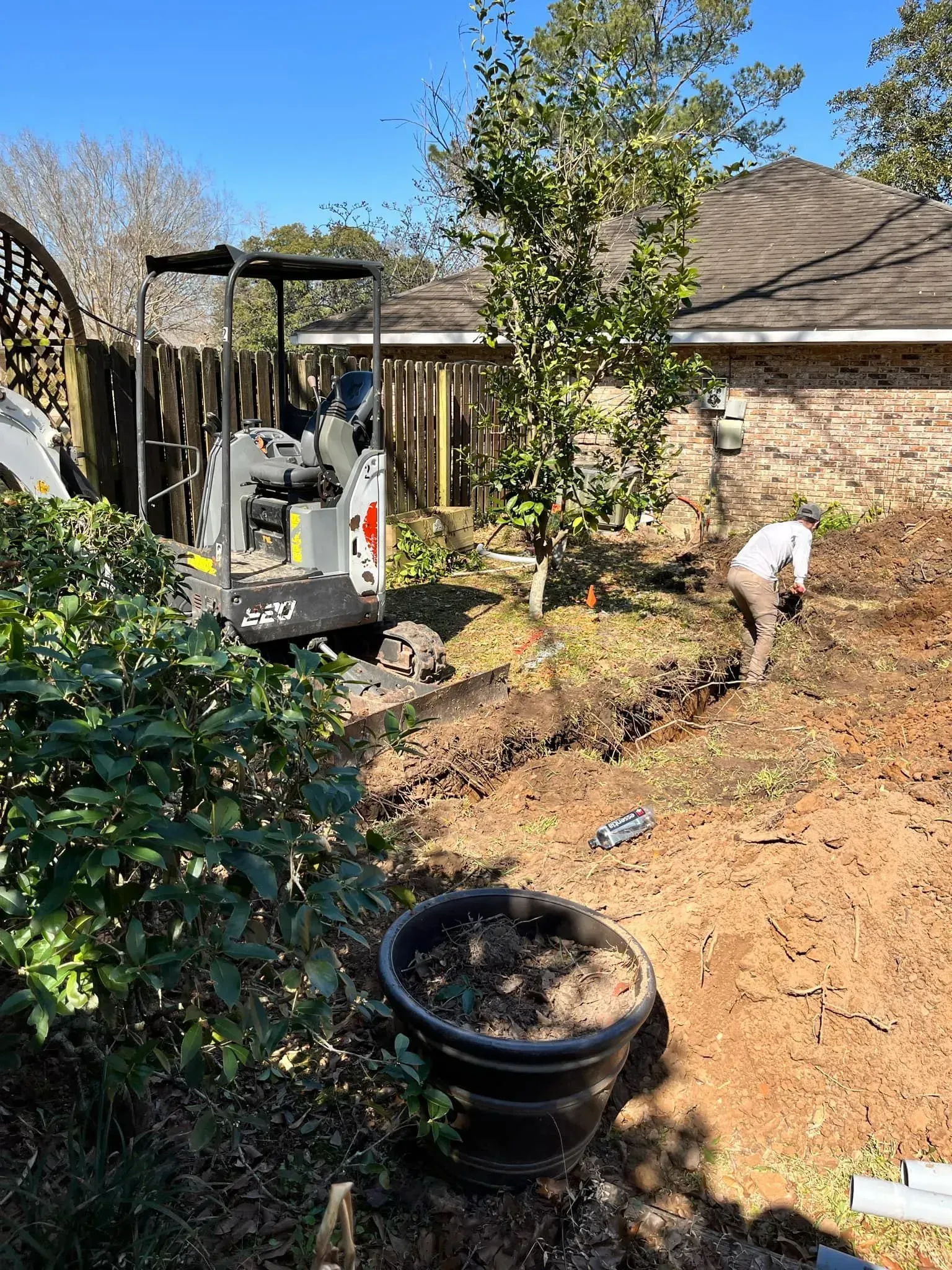 Backyard with mini excavator and worker digging a trench, next to foliage, in sunlight.