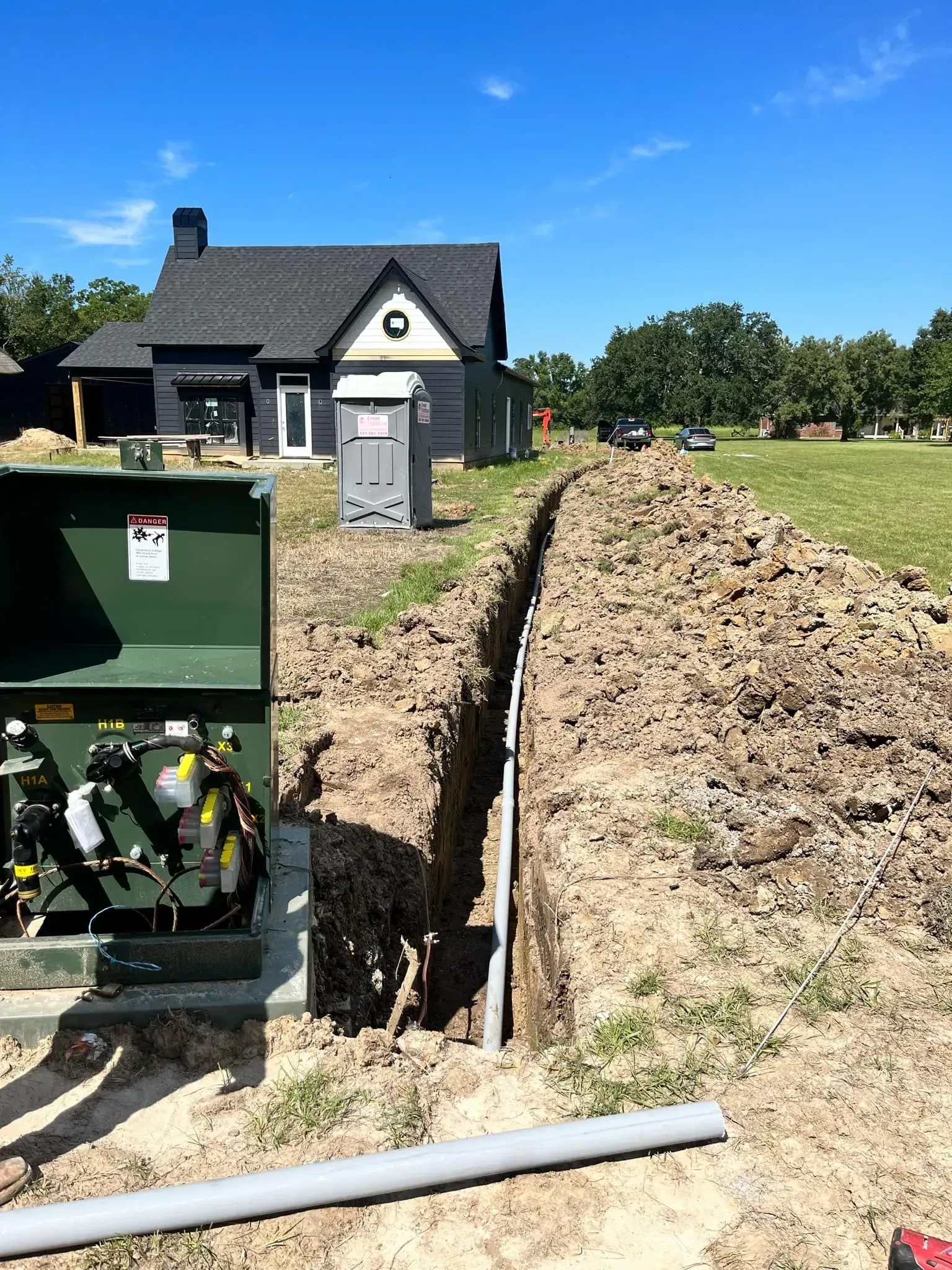 Green electrical box and trench with conduit leading to a dark-colored house on a sunny day.