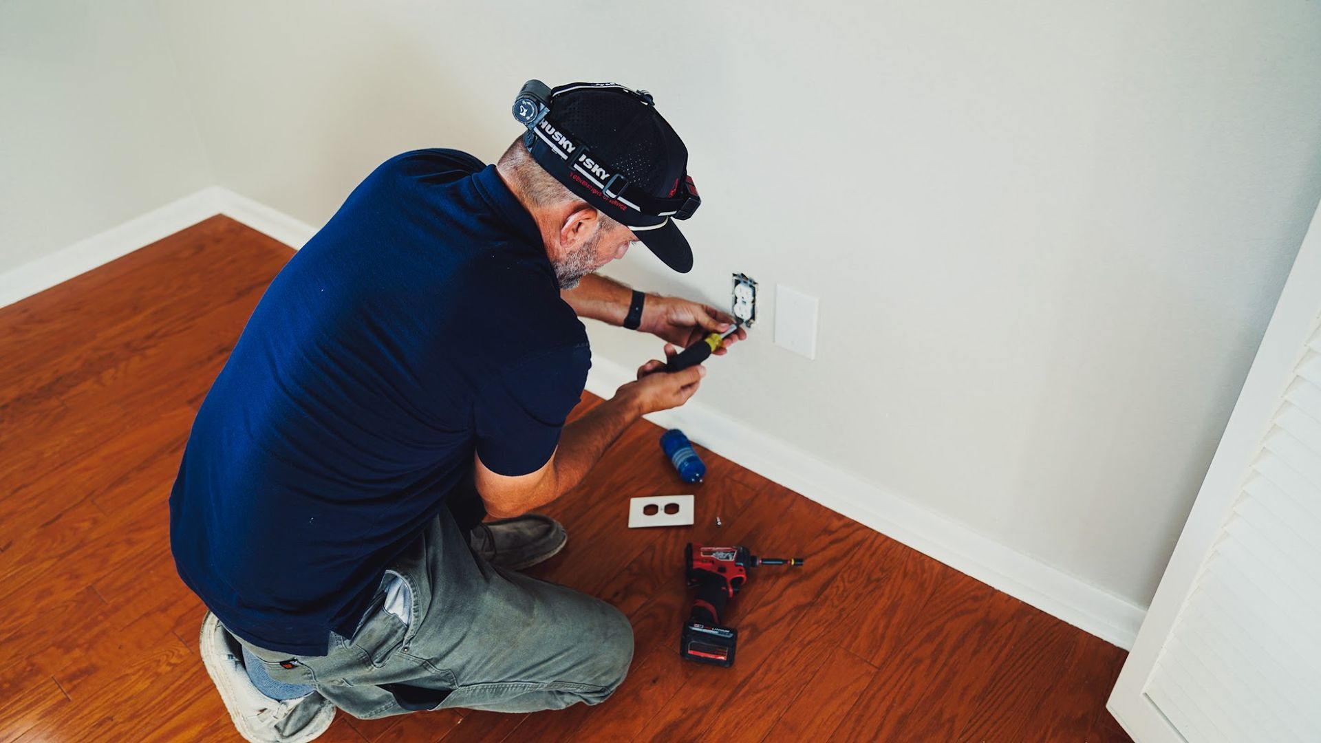 Man kneeling on a wood floor installing an electrical outlet on a white wall.