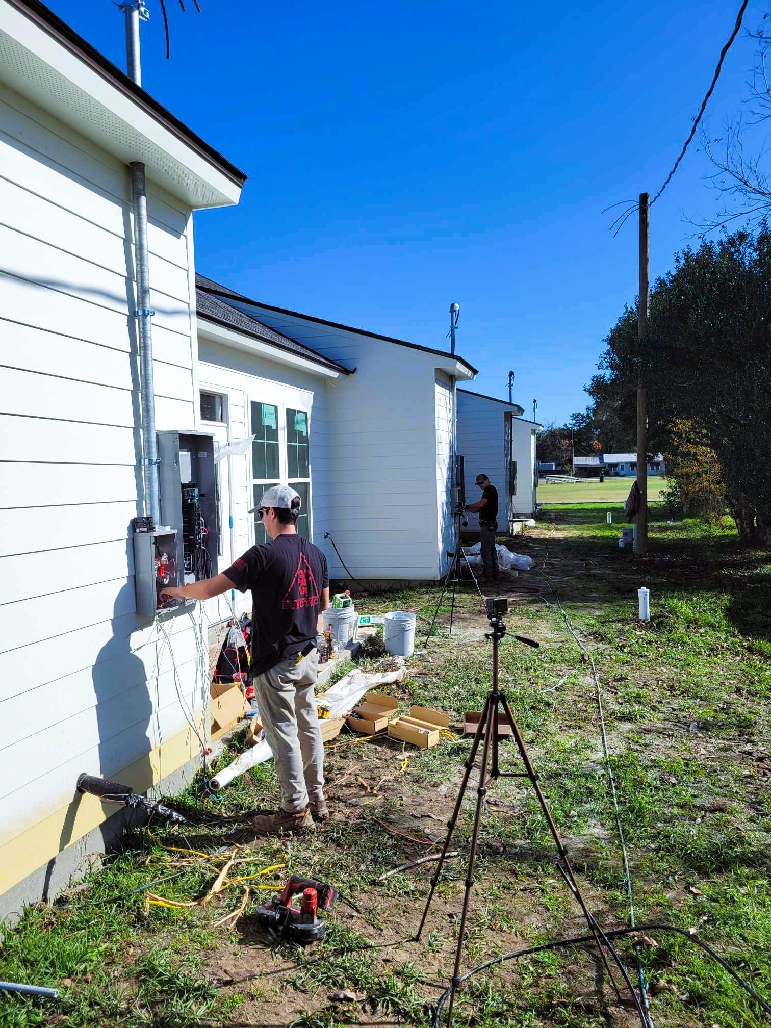 Two workers installing electrical panels on the side of white buildings under a blue sky.