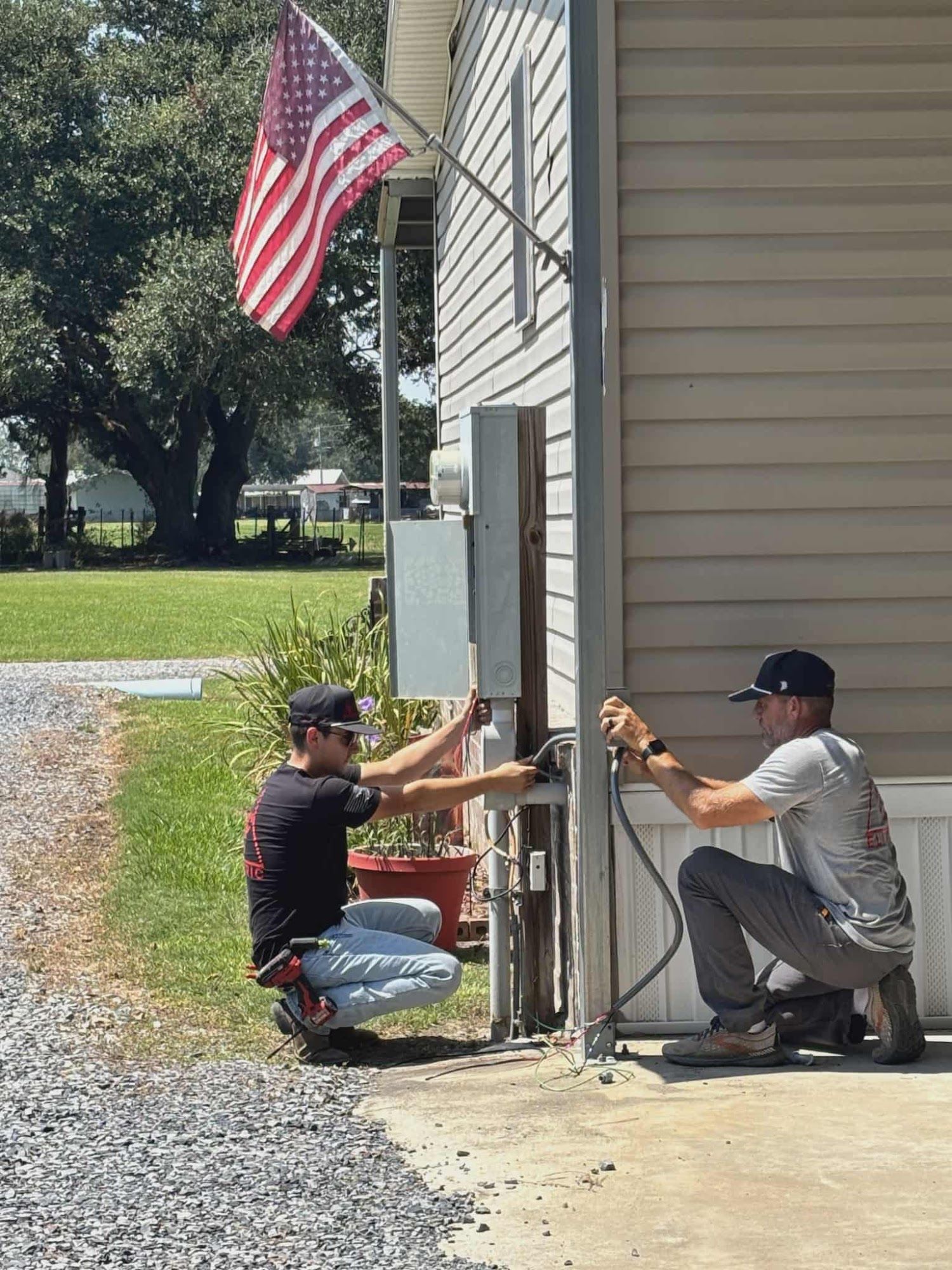 Two people installing electrical equipment on a house exterior; American flag in the background.