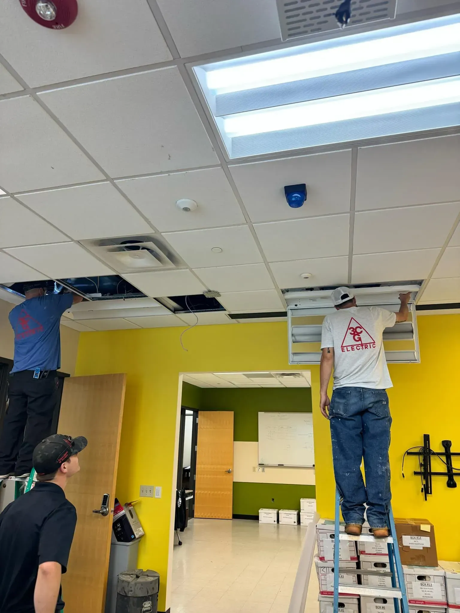 Three workers installing ceiling lights in a room with a lime green wall, white ceiling tiles, and a yellow doorway.