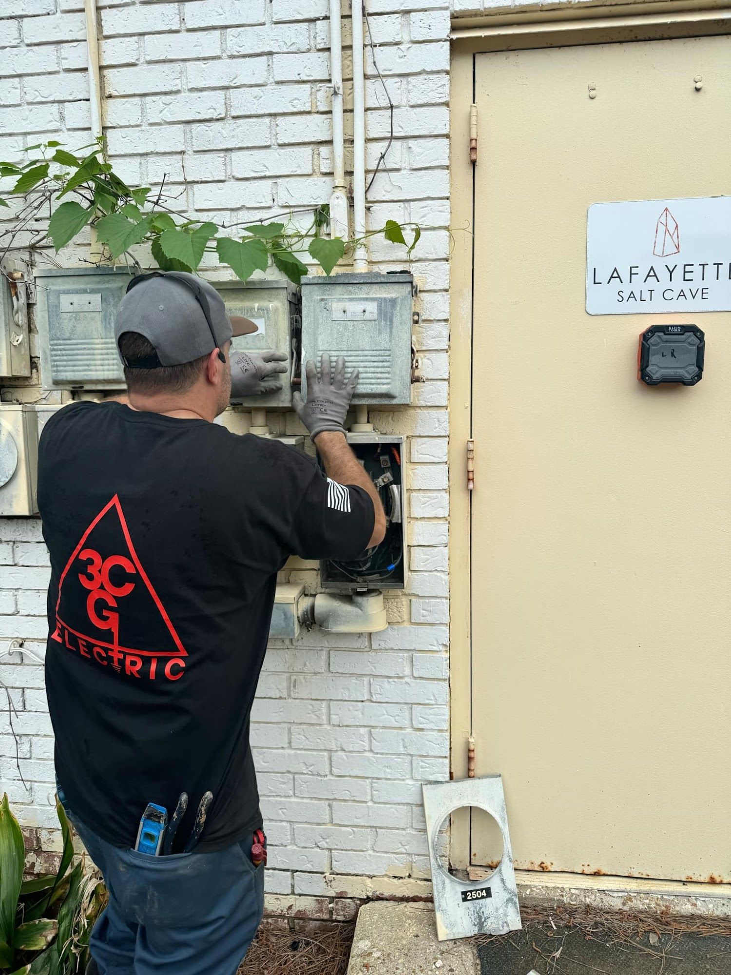 Electrician working on electrical meters mounted on a brick wall, near a beige door with a sign.