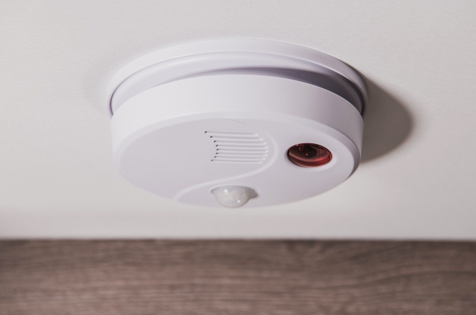 White smoke detector mounted on a white ceiling, with a visible light and speaker.